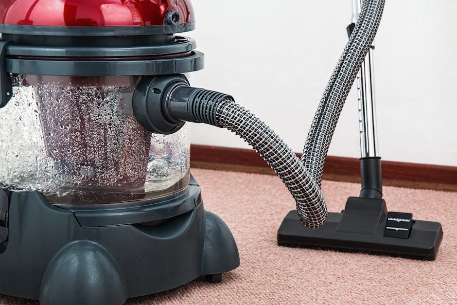 A woman engaged in surface cleaning using a yellow vacuum cleaner with a black hose attachment, positioned on a patterned, light-colored rug with floral motifs. The room features a wooden floor visibl