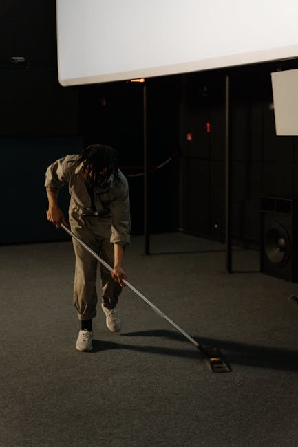 A person cleaning a dark grey carpeted floor in a dimly lit room, using a long-handled tool for surface cleaning. The room features a large white projection screen on the wall, black partition panels,