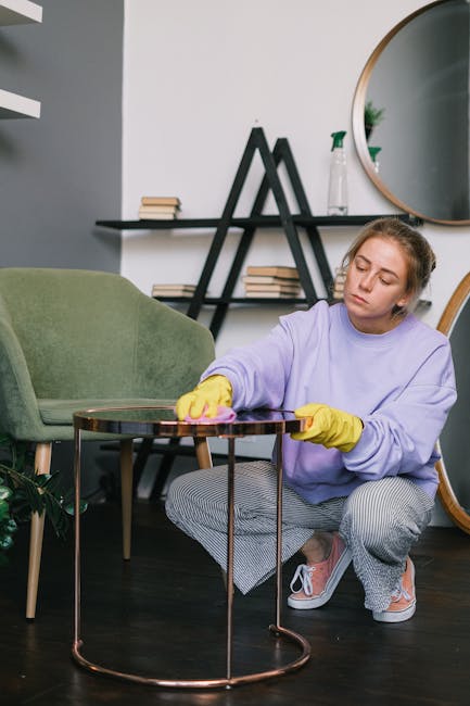 Close-up of a person in a dark jacket using a handheld vacuum cleaner to deep clean a yellow and white patterned cushion placed on a dark fabric sofa, demonstrating upholstery surface cleaning in a we